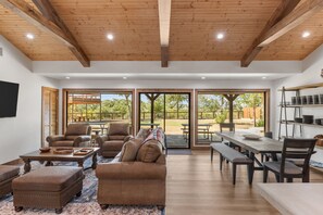 A spacious living area with a wooden ceiling, large sliding glass doors leading to an outdoor patio, and a dining table set for four. - Guadalupe Bluff Trail House 5 Bedroom Home (Kerrville)