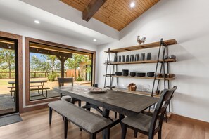 Un coin repas avec une table et des bancs en bois, une étagère en métal où sont rangés de la vaisselle, et une vue sur l’extérieur.