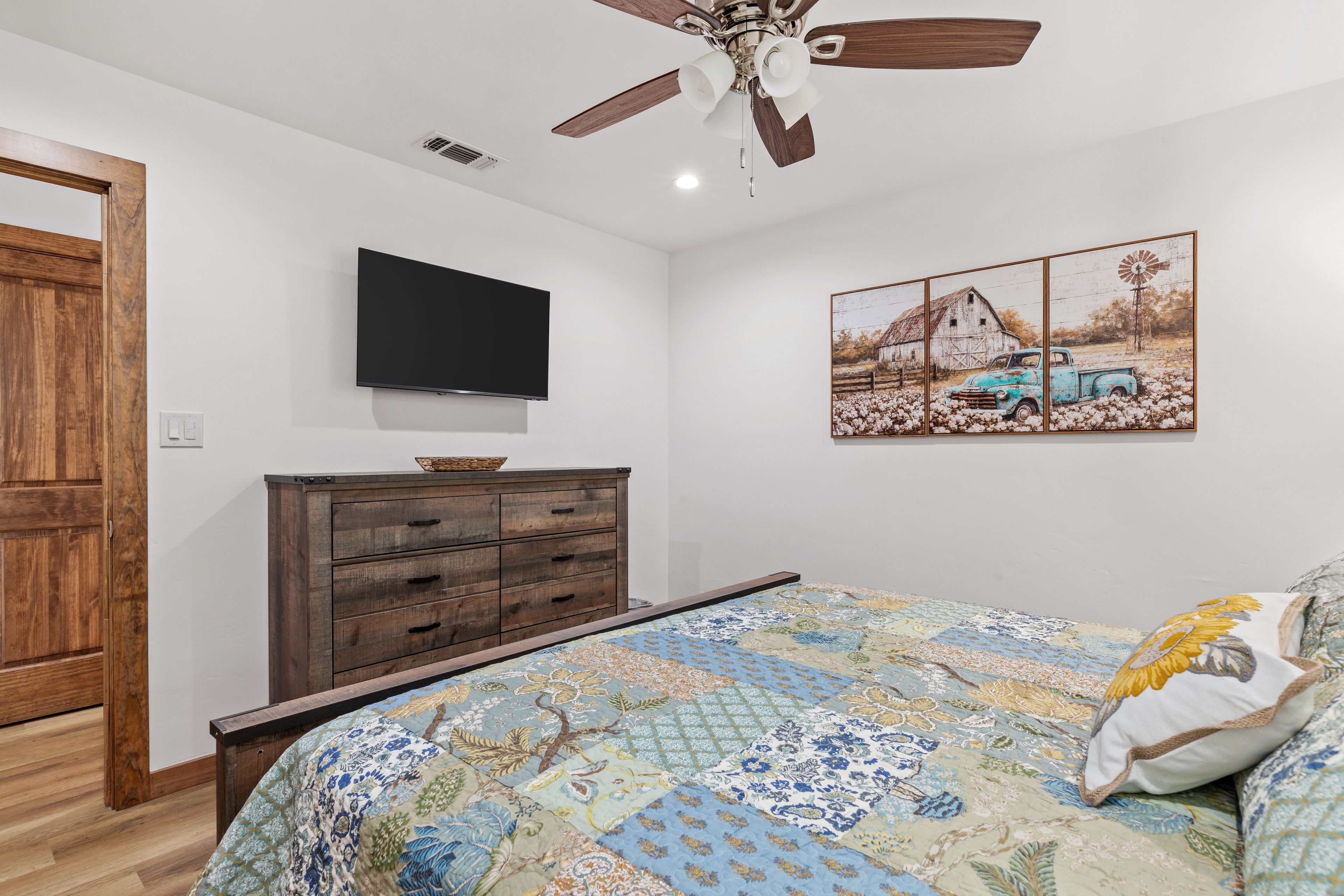 A bedroom with a bed, a wooden dresser, a ceiling fan, a flat-screen TV, and a painting of a barn and truck.