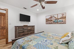 A bedroom with a bed, a wooden dresser, a ceiling fan, a flat-screen TV, and a painting of a barn and truck. - Guadalupe Bluff Trail House 5 Bedroom Home (Kerrville)