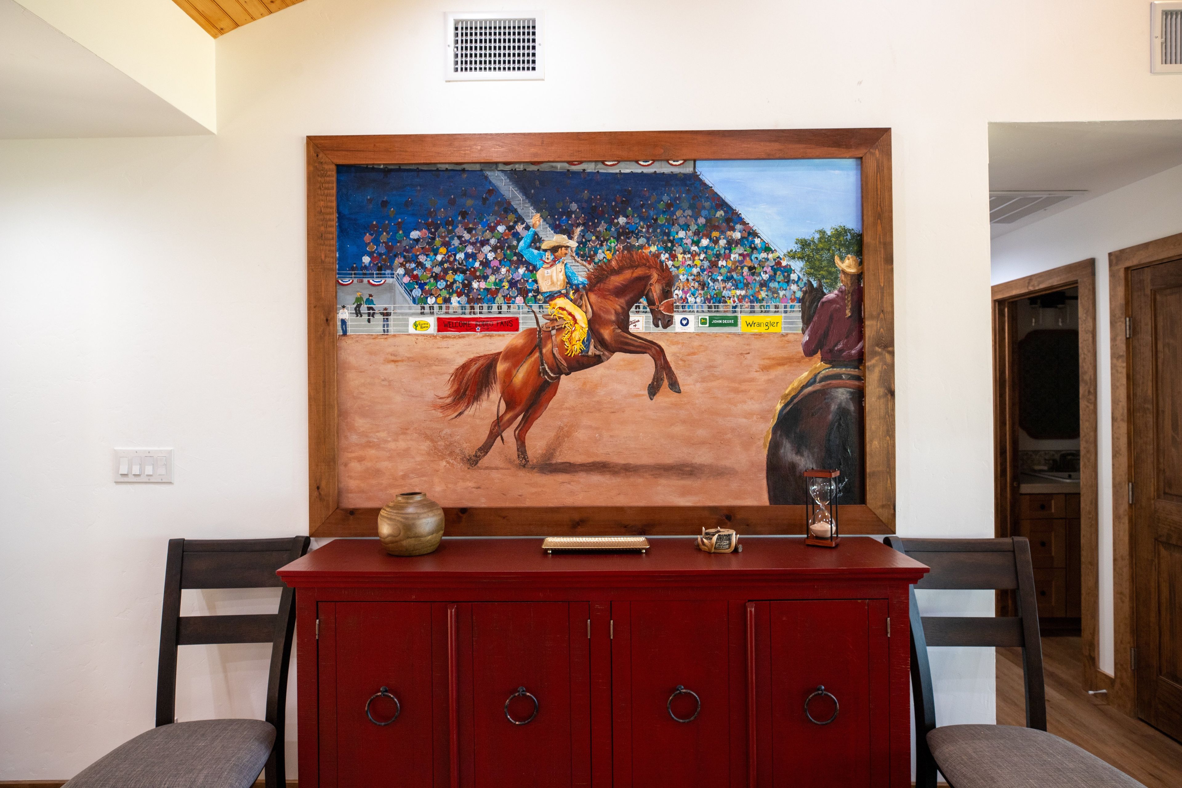 A painting of a rodeo scene on a wall above a red cabinet with drawers.