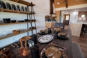 A kitchen with wooden shelves holding various bowls and plates, a dining table with cutlery, and a kitchen island with a coffee machine. - Guadalupe Bluff Trail House 5 Bedroom Home (Kerrville)