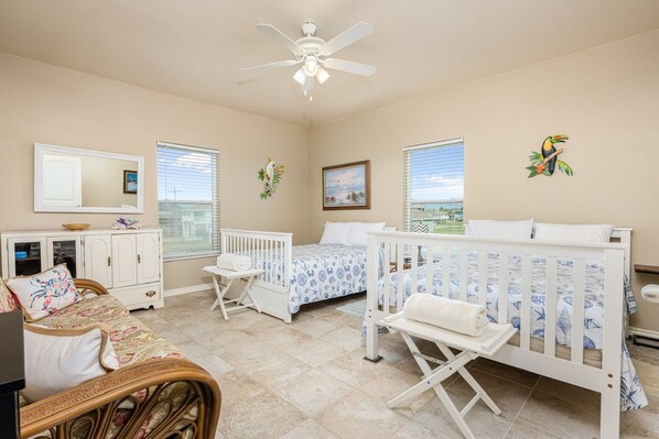 A bedroom with a crib, a bed with a blue and white patterned bedspread, a white dresser, a wicker sofa, and a ceiling fan. - Shore Enough 3 Bedroom Home (Port Bolivar)