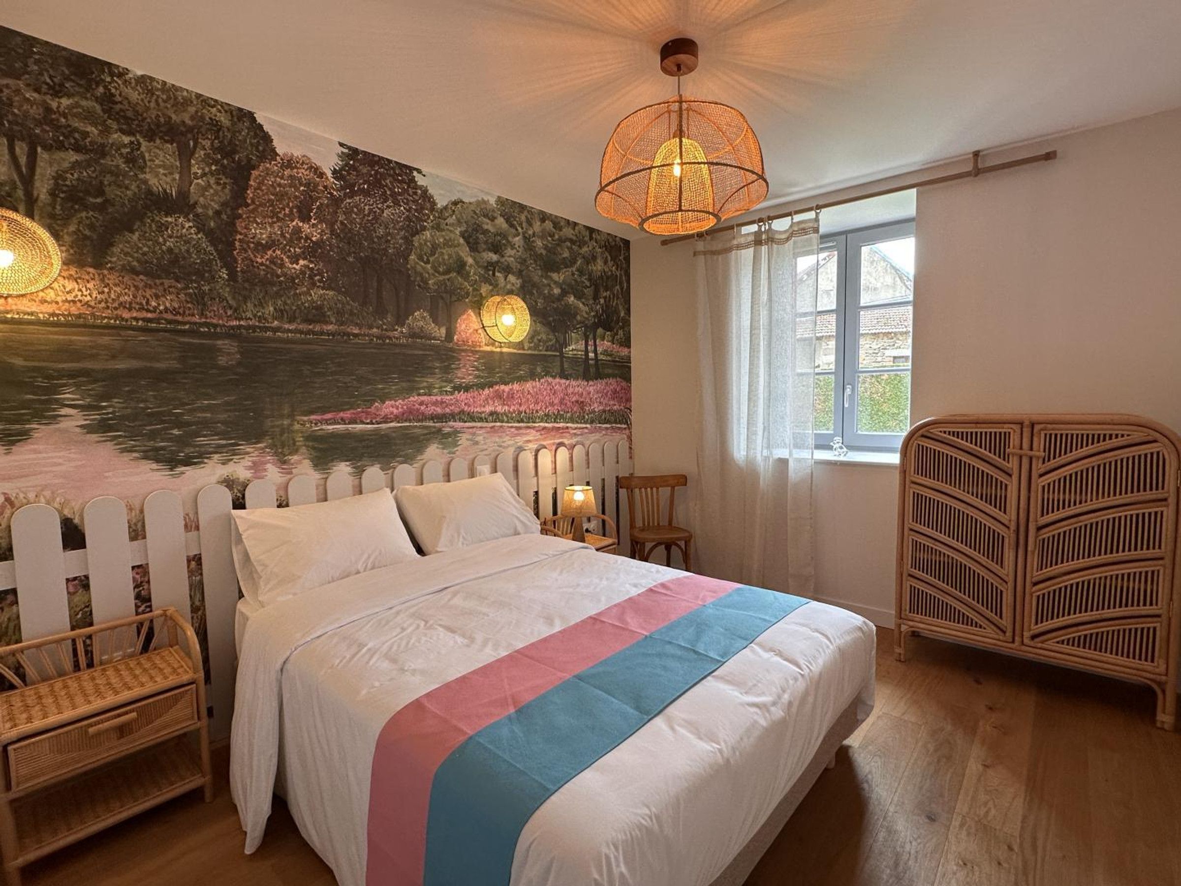 Quarto com uma cama grande, um mural na parede, uma janela e uma cadeira de vime.