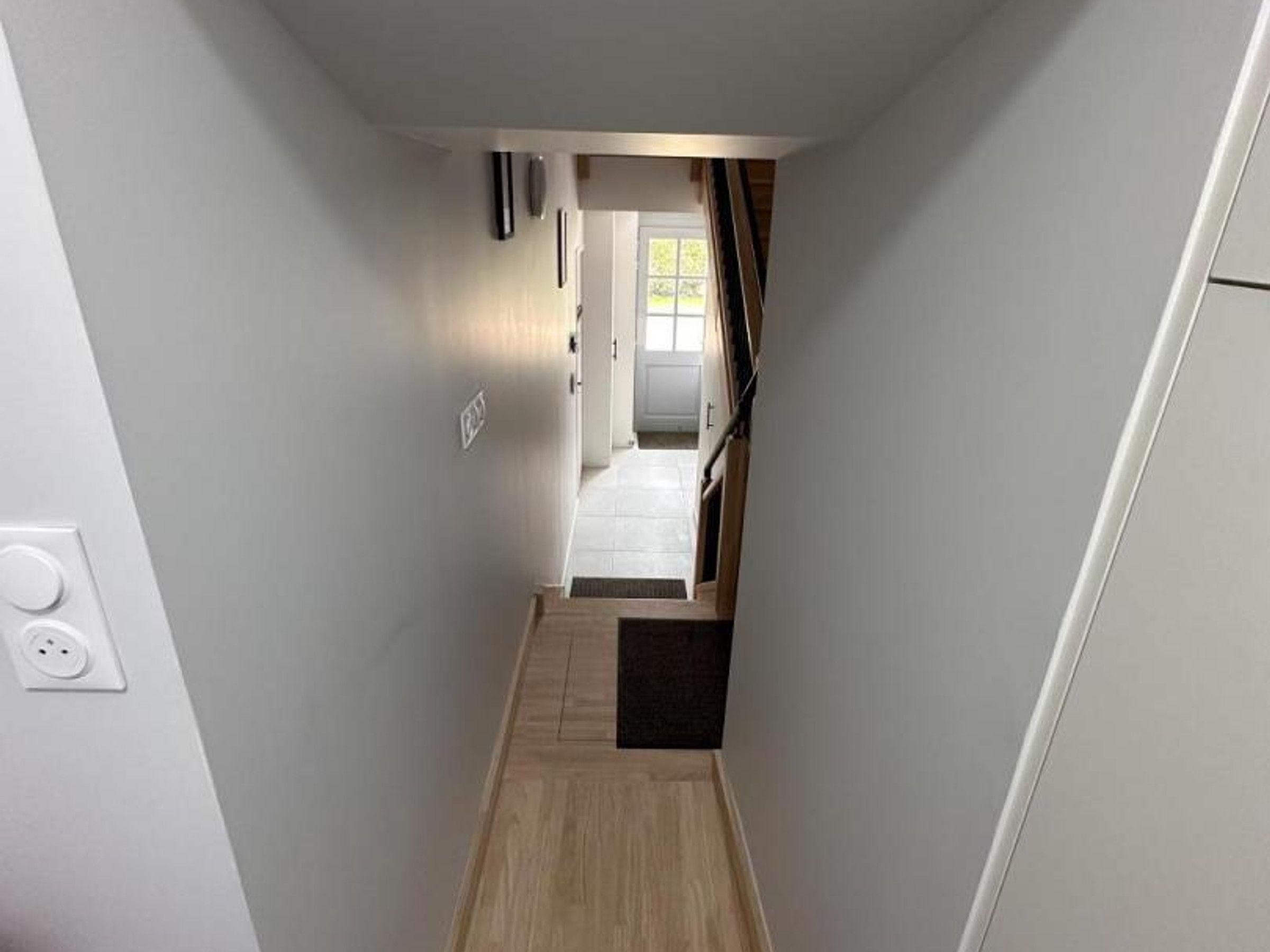 Een smalle gang met een houten vloer die leidt naar een kamer met een deur en een raam.