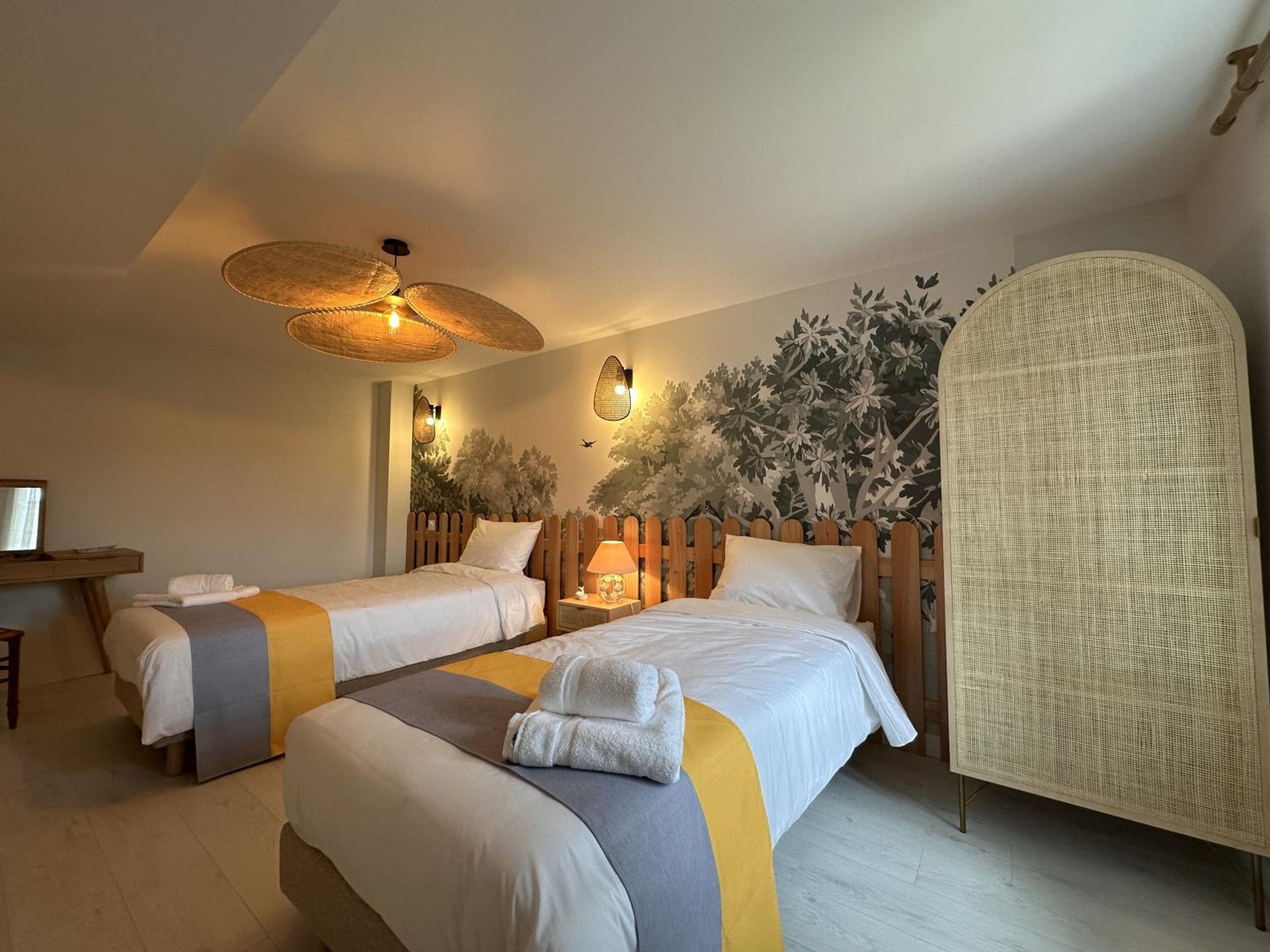 Una habitación de hotel con dos camas, un cabecero de madera, una silla de mimbre y un ventilador de techo.