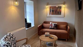A cozy living room with a brown sofa, a patterned armchair, a small round table with a book and remote, and a wall-mounted artwork.
