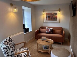 A cozy living room with a brown sofa, a patterned armchair, a small round table with a book and remote, and a wall-mounted artwork.