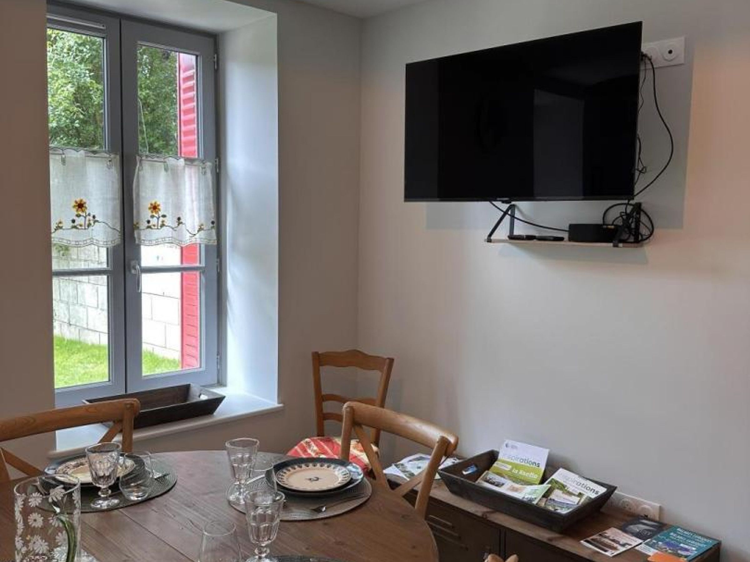 A dining area with a table set for two, a television mounted on the wall, and a window with curtains.