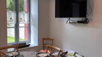 A dining area with a table set for two, a television mounted on the wall, and a window with curtains.