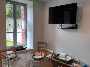 A dining area with a table set for two, a television mounted on the wall, and a window with curtains.