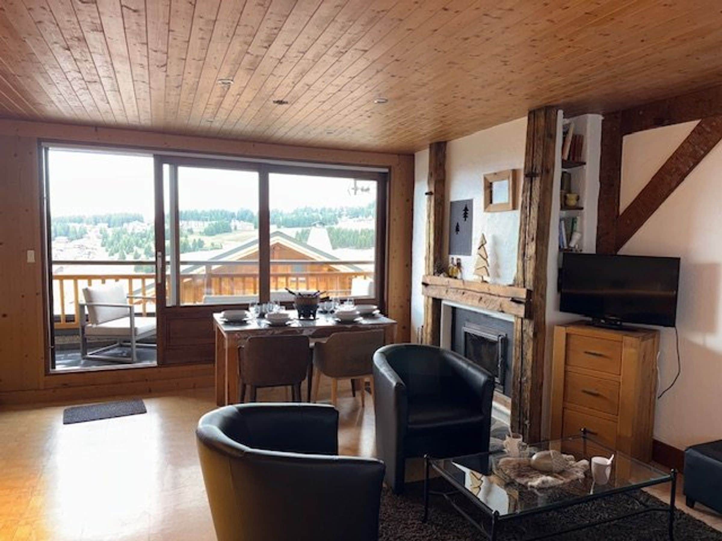 Een ruime woonkamer met een eettafel, stoelen, een open haard, een televisie en een groot raam met uitzicht op gebouwen en bomen.