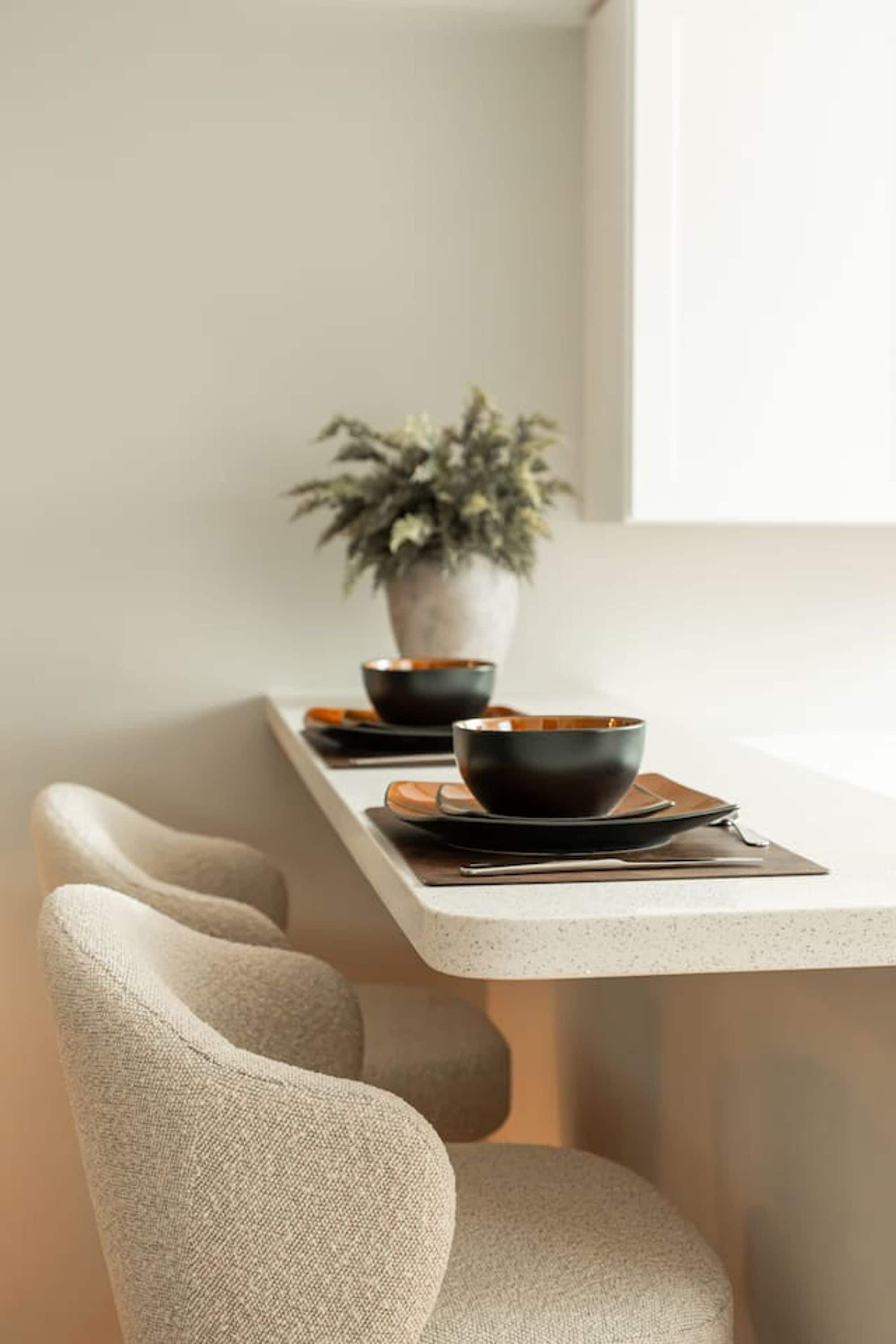 A minimalist dining area with a long table, two black bowls, a potted plant, and a window.
