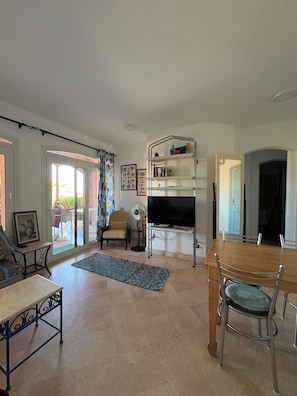 A living room with a television on a stand, a dining table with chairs, a sofa, a chair, a fan, and a rug. - 2BR Apartment in West Golf, El Gouna (Red Sea Governorate)
