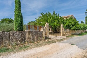 A rural property with a gravel driveway leading to a gated entrance, surrounded by stone walls and lush greenery. - La Maison Rouge Garance (Saint-Hilaire-d'Ozilhan)