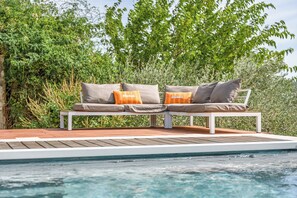 A poolside seating area with a bench and cushions, surrounded by greenery. - La Maison Rouge Garance (Saint-Hilaire-d'Ozilhan)