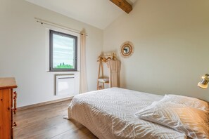 A bedroom with a bed, a window, a wooden dresser, a mirror, and a wall-mounted light fixture. - La Maison Rouge Garance (Saint-Hilaire-d'Ozilhan)