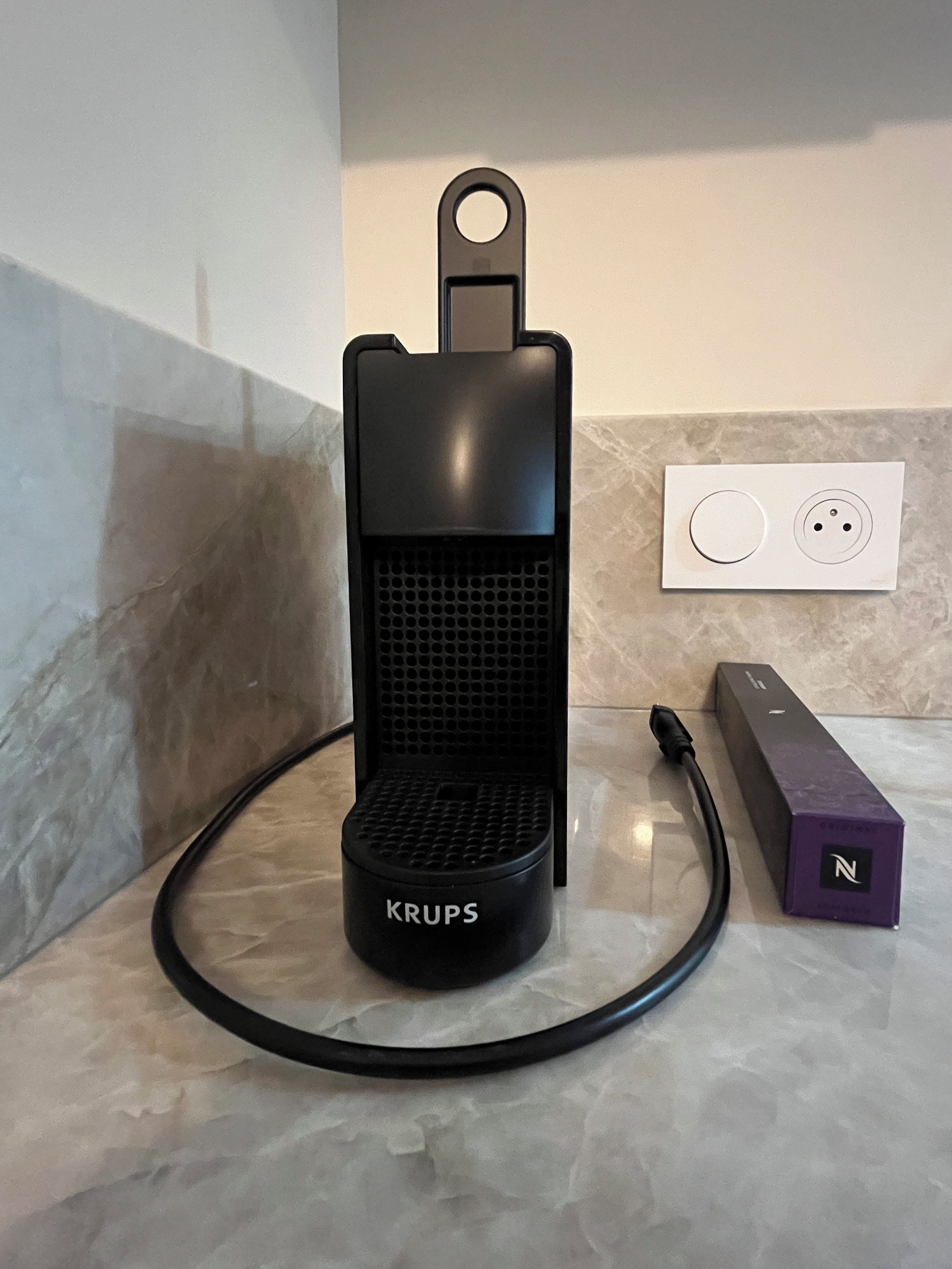 A Krups coffee maker on a marble countertop with a power cord and a purple device beside it.