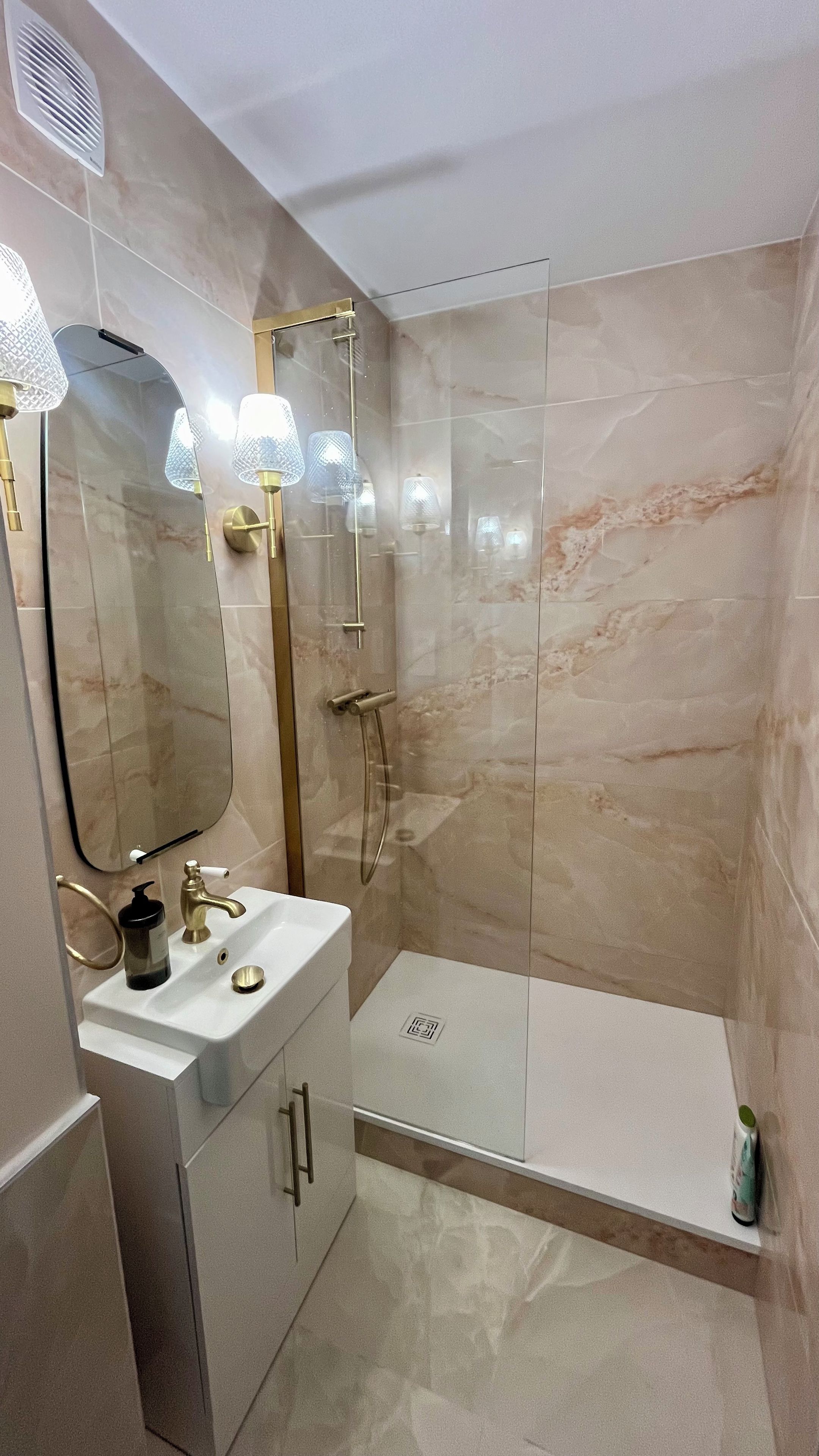 Ein Badezimmer mit einer Duschkabine, einem weißen Waschbecken mit goldfarbenen Armaturen und einem großen Spiegel mit Pendelleuchten.