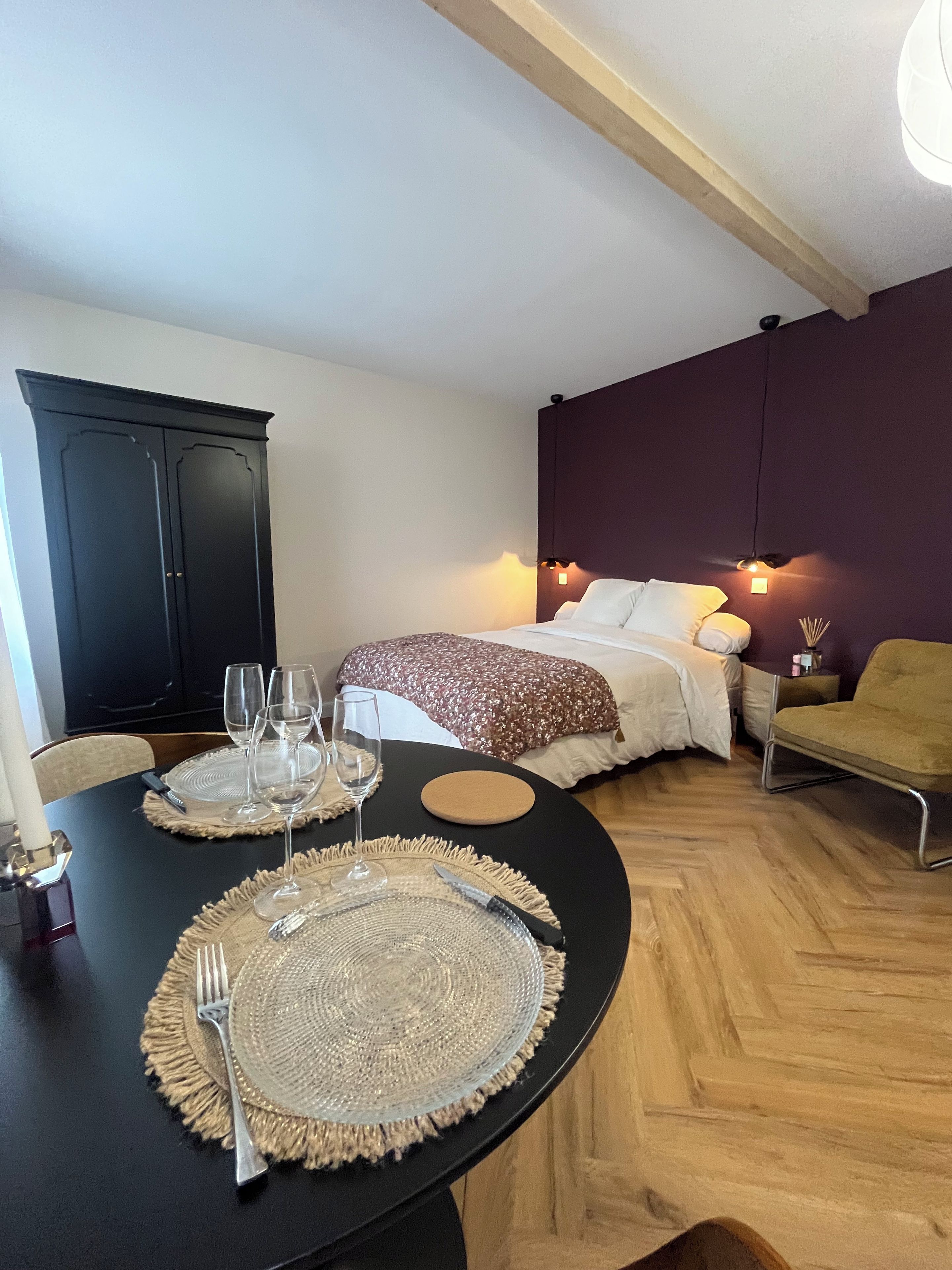 A room with a dining table set for two, a bed with a headboard, a chair, and a wardrobe.