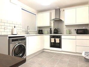 A compact kitchen with a washing machine, oven, microwave, and a gas stove. - Cosy 2-Bed | 3 Space Parking | M56 Nearby (Helsby)