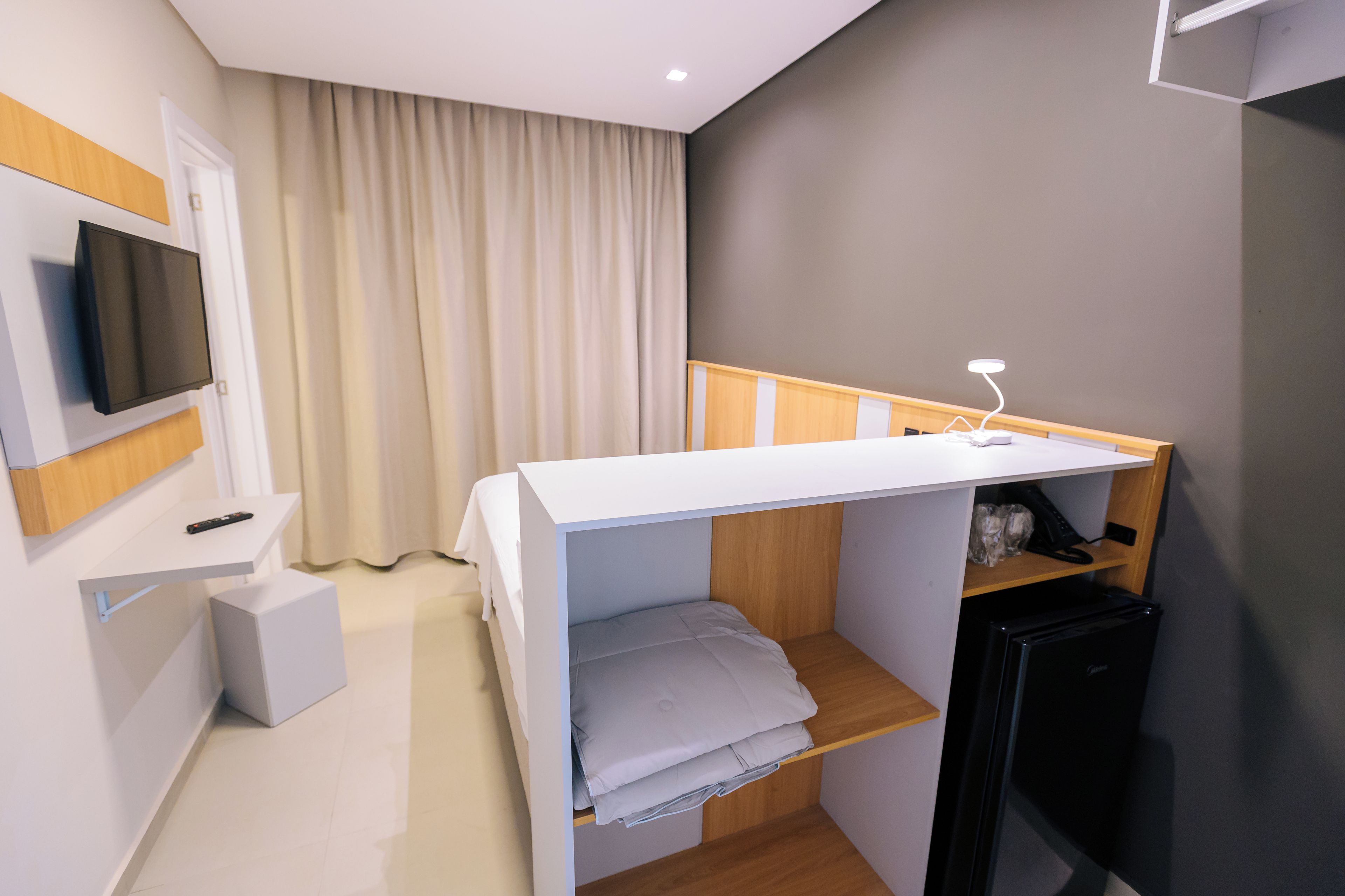 Ein kleines Hotelzimmer mit Etagenbett, Schreibtisch mit Stuhl, Wandfernseher, kleinem Kühlschrank und Nachttisch.