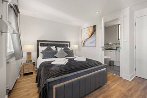 A modern bedroom with a large bed, bedside tables, a nightstand, a painting, and a view into the bathroom. - Elegant 3bed Residence Marylebone & Paddington (Greater London)