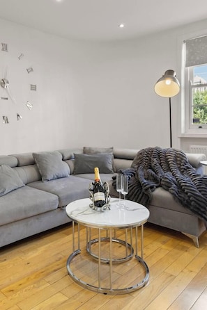 A modern living room with a grey sofa, a round coffee table with a bottle and glasses, and a wall clock. - Elegant 3bed Residence Marylebone & Paddington (Greater London)
