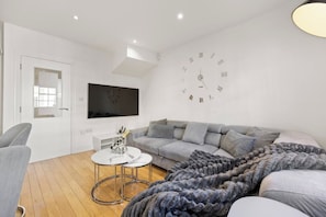 A modern living room with a grey sofa, a round coffee table, a flat-screen TV mounted on the wall, and a large wall clock. - Elegant 3bed Residence Marylebone & Paddington (Greater London)