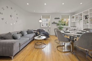 A modern living room with a grey sofa, a coffee table, and a dining area with chairs and a table set for a meal. - Elegant 3bed Residence Marylebone & Paddington (Greater London)