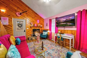 Una sala de estar acogedora con chimenea, un sofá con cojines de colores, un escritorio con computadora y una pantalla de televisión grande.