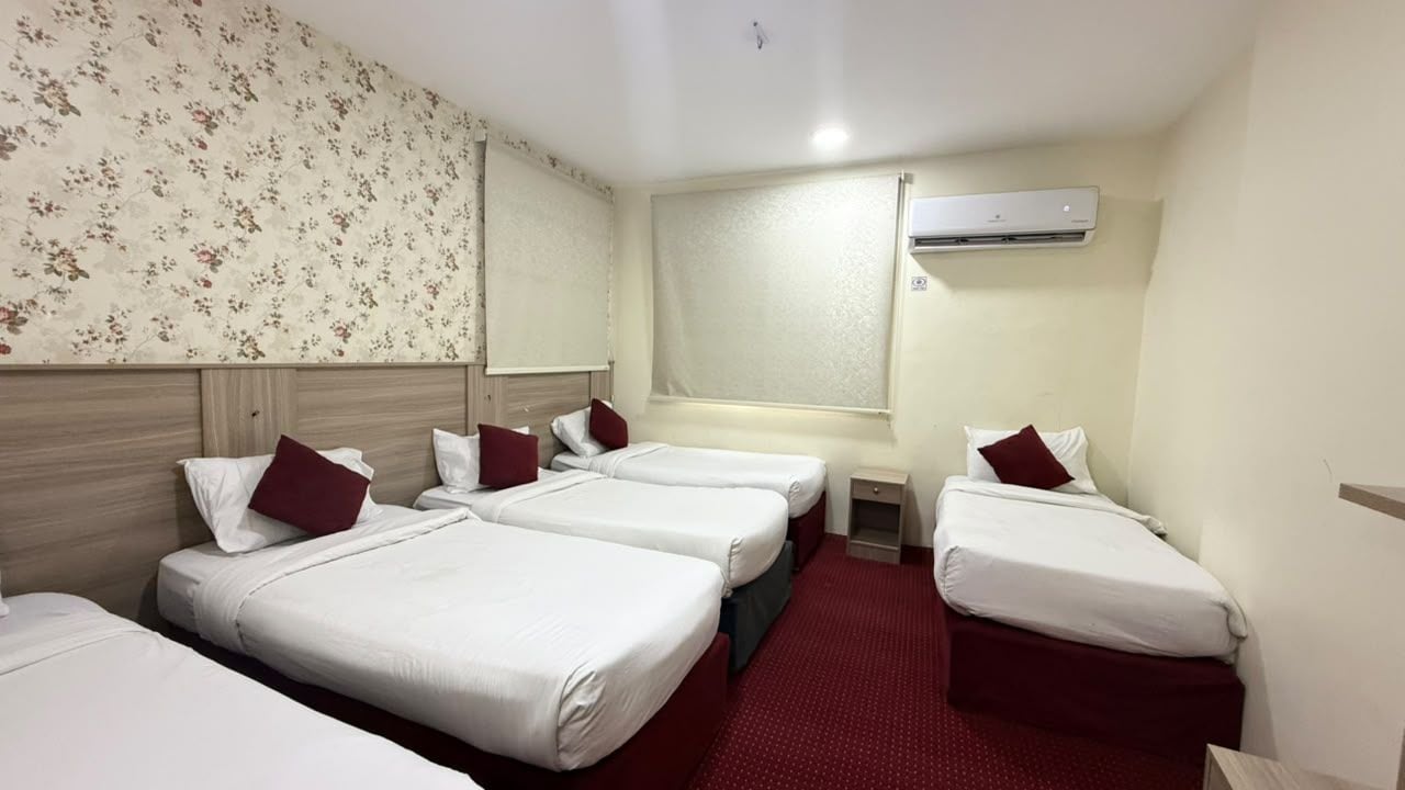 A hotel room with four single beds, a small bedside table, a red carpet, and an air conditioning unit on the wall.