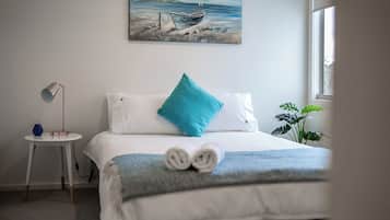 Ein ordentlich bezogenes Bett mit einem blauen Kissen und einem zusammengerollten Handtuch, ein kleiner Nachttisch mit einer Lampe, ein Bild eines Bootes an der Wand und ein Fenster mit einer Pflanze daneben.