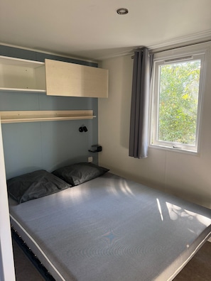 Habitació petita amb un llit individual, escriptori plegable, finestra amb cortines i prestatge sobre el llit.