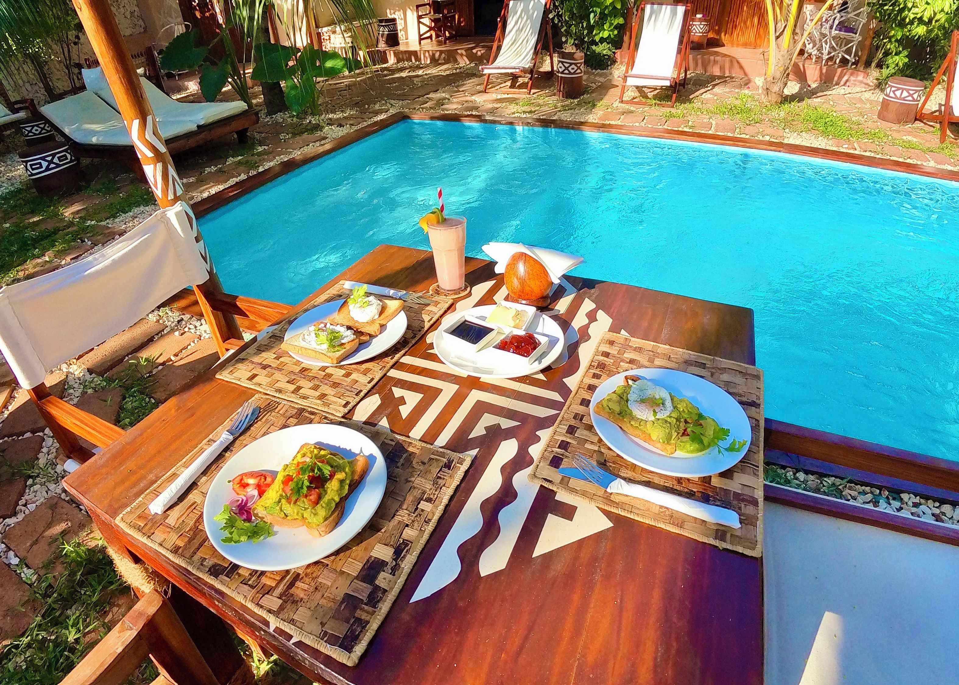 Zona de comedor junto a la piscina, con una mesa preparada para dos, mostrando diversos platos y una bebida.