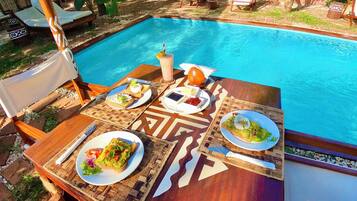 Zona de comedor junto a la piscina, con una mesa preparada para dos, mostrando diversos platos y una bebida.