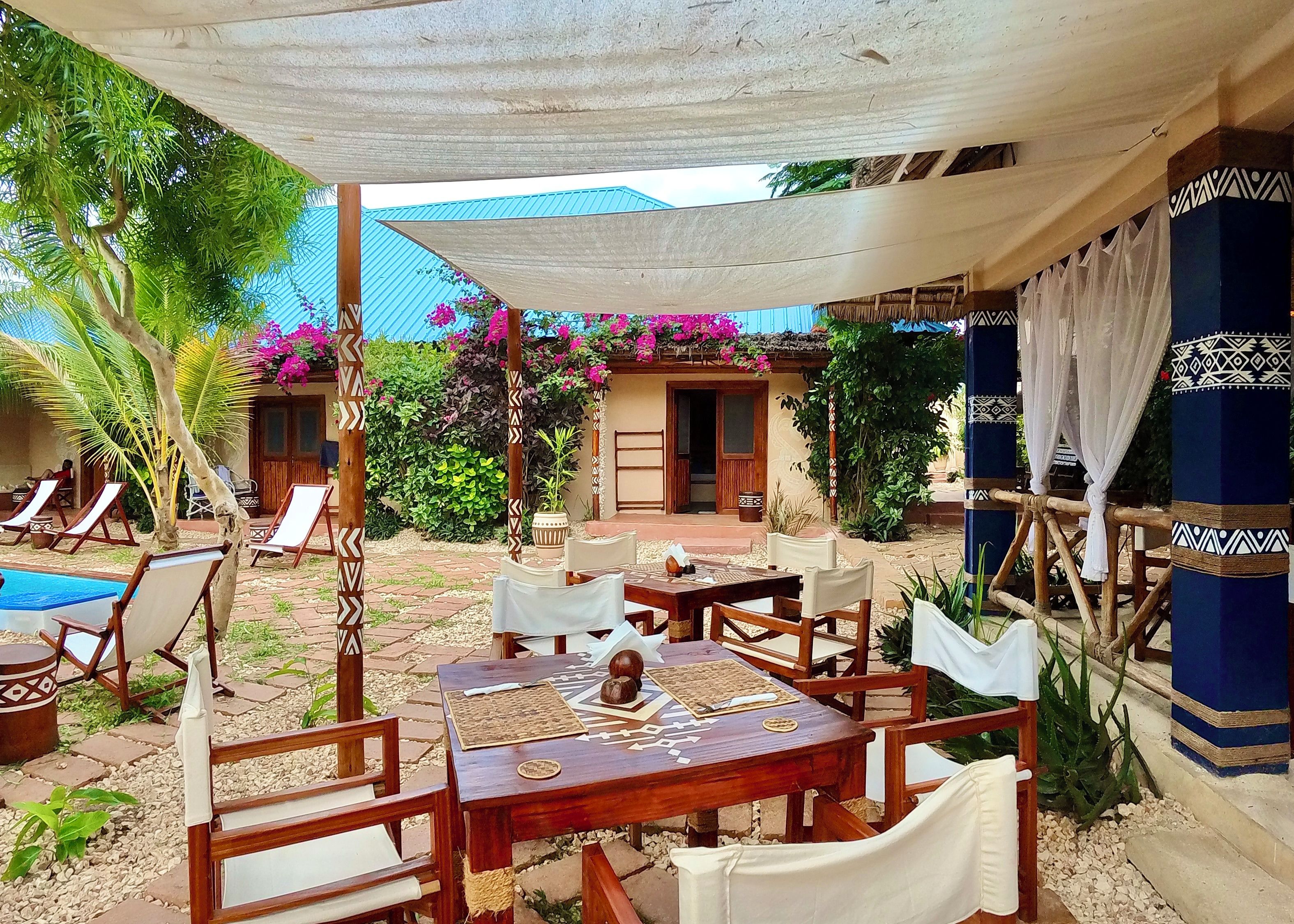 Un patio exterior con mesa y sillas, rodeado de una zona sombreada, una piscina y un edificio con techo azul.
