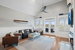A spacious living room with a high ceiling, a ceiling fan, a fireplace, and a view of the outdoors through large windows. - Sugar Sand Cottage at Destin Pointe (Destin)