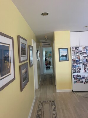 A hallway with framed pictures on the walls, a rug on the floor, and a refrigerator with photos on the right. - The Scotch Bonnett Beach House - Best Location! (Nags Head)