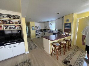 A kitchen with a central island, bar stools, a refrigerator with photos, and a television on a stand. - The Scotch Bonnett Beach House - Best Location! (Nags Head)
