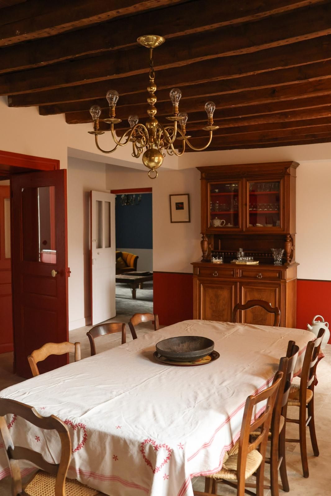 Uma sala de jantar com um lustre, cadeiras de madeira, uma mesa com toalha branca e um aparador de madeira com portas de vidro.