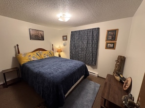 A bedroom with a bed, a nightstand, a lamp, a window with curtains, and a wooden dresser.