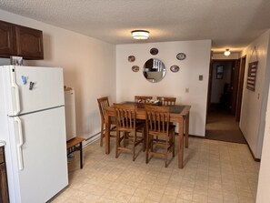 Une cuisine avec un réfrigérateur blanc, une table à manger en bois et des chaises. On y trouve une petite table sur laquelle est posé un panier. Les murs sont décorés d’assiettes et d’un miroir.