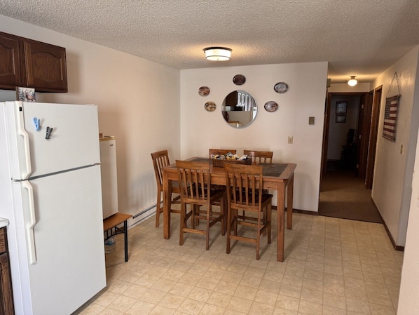 A kitchen with a white refrigerator, wooden dining table, and chairs. There is a small table with a basket on it. The walls have decorative plates and a mirror.