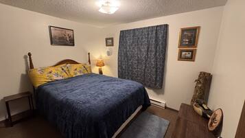 Ein Schlafzimmer mit Bett, Nachttisch, Lampe, einem Fenster mit Vorhängen und einer Holzkommode.