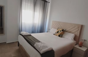Ein Hotelzimmer mit Bett, Nachttisch, einem Fenster mit Vorhängen und einem an der Wand montierten Fernseher.