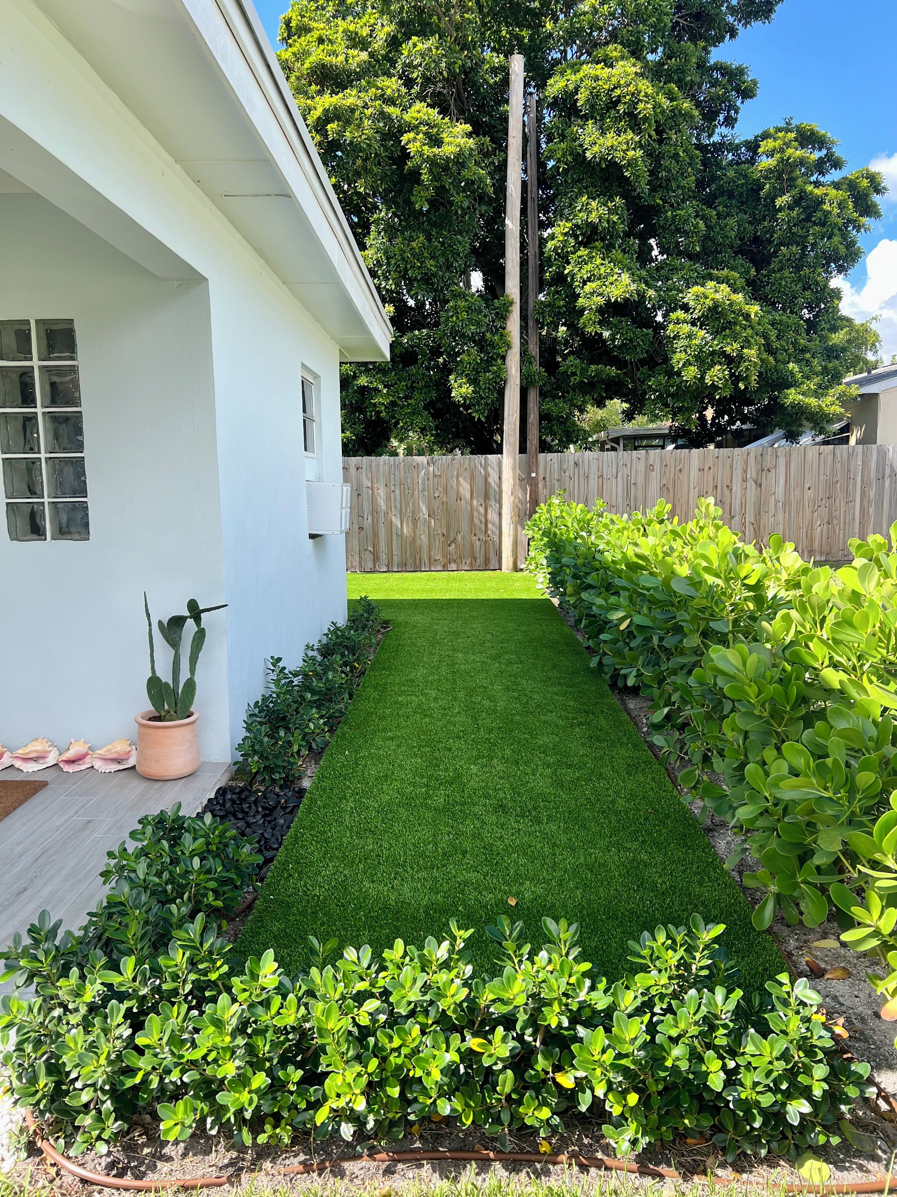 Un jardin à l’arrière d’une maison, avec une maison blanche, une clôture en bois et diverses plantes vertes.