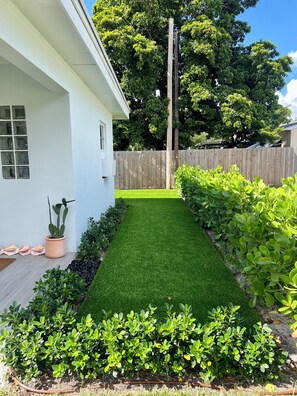 A residential backyard with a white house, a wooden fence, and a variety of green plants.