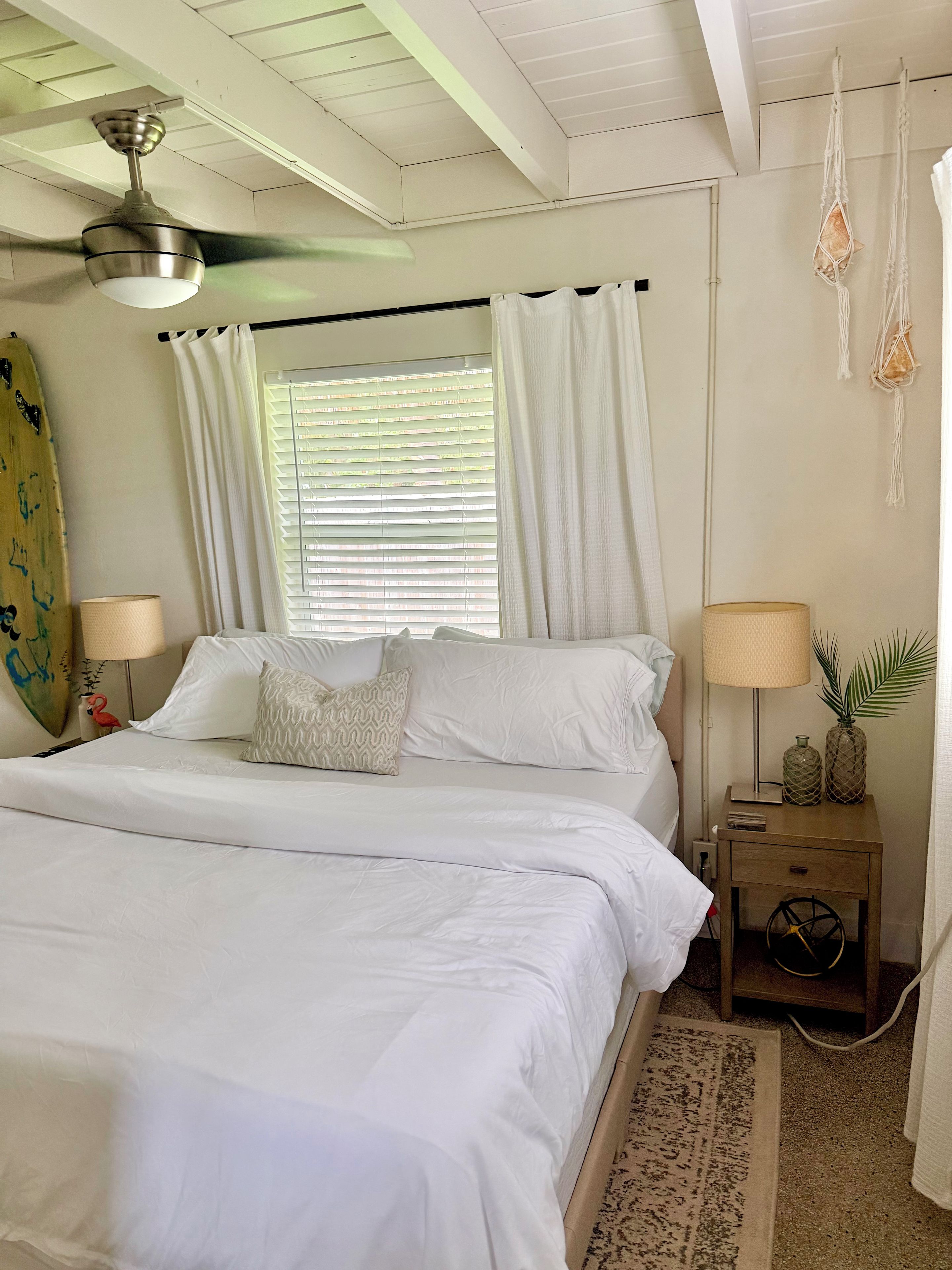 A bedroom with a bed, two bedside tables, a ceiling fan, a window with curtains, and a surfboard on the wall.