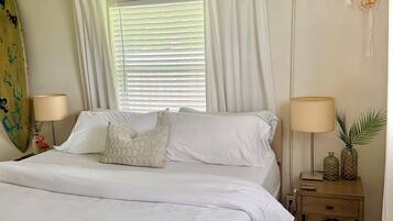 A bedroom with a bed, two bedside tables, a ceiling fan, a window with curtains, and a surfboard on the wall.